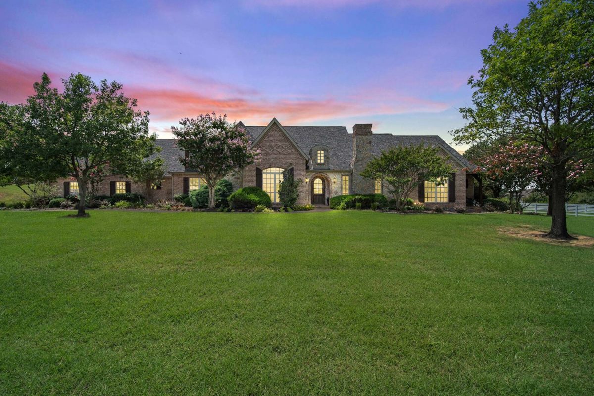 Brick house across a wide lawn at dusk, with warm lit windows under a pink and violet sky